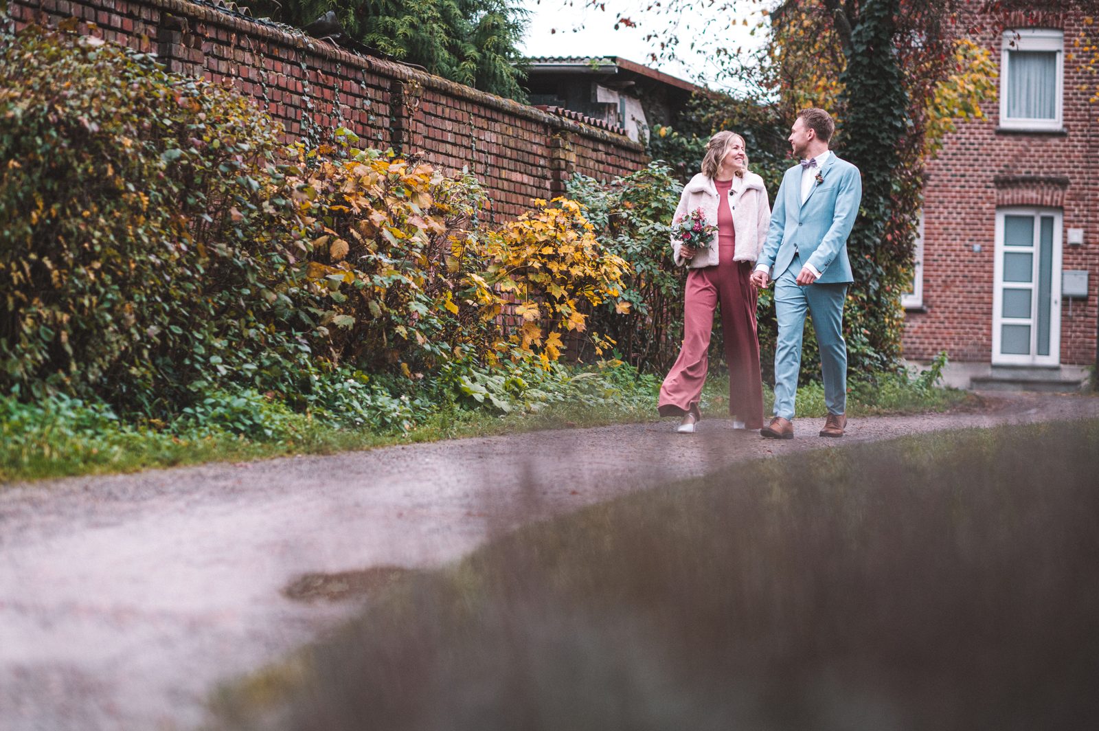 Brautpaar hält lachend Hände auf einem herbstlichen Weg zwischen altem Backsteinmauerwerk – Hochzeitsfotografie von Sascha Uhle im Münsterland